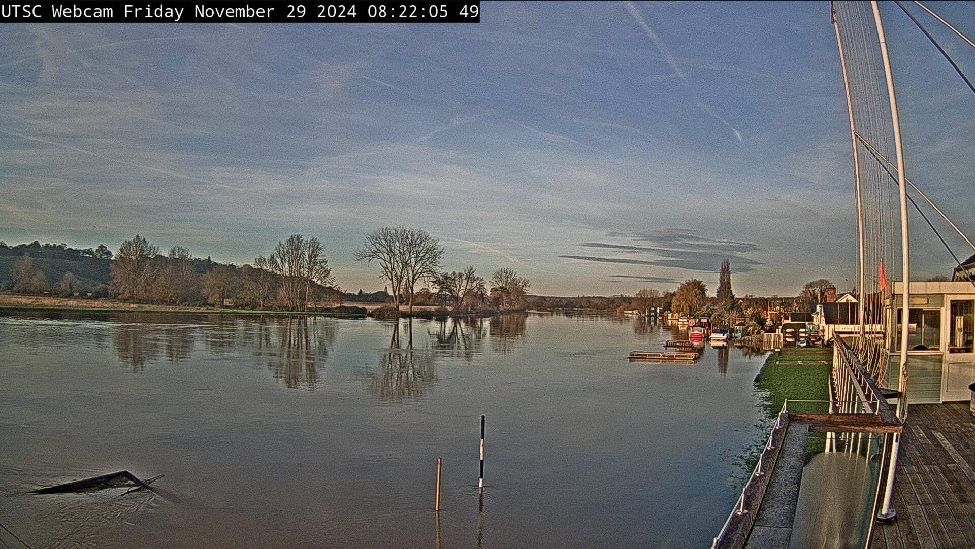 A bright Autumn morning looking over the River Thames in flood. The water has risen about 2 ft and is now covering the Thames Path outside the Upper Thames sailing club. The water has a harmless glassy look and looks calm even though the water is racing beneath the surface. The trees on the opposite bank are reflected on the water&apos;s surface, but the pontoons next to the bank now look like they are in the middle of the river.