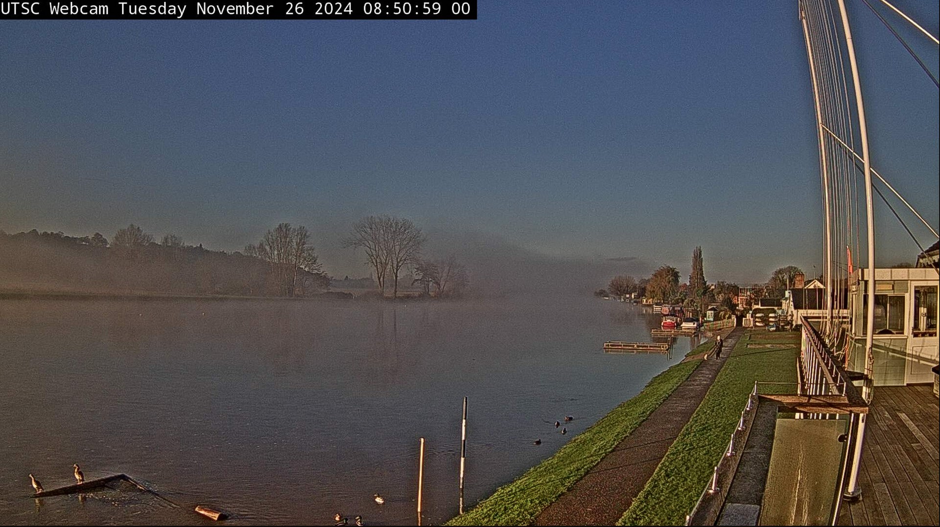 A dark grey cloud looms over the River Thames, yet the rest of the sky is bright blue. The water is rising up the bank on this Tuesday morning and the black foot marker is half covered.